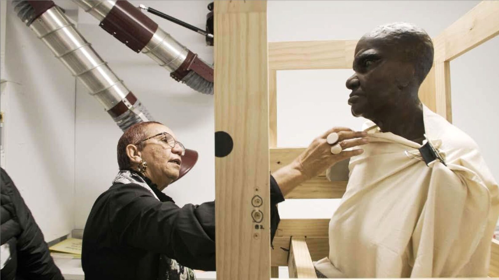 Her Name Is Nanny Nellie — a woman reaches towards a sculpted bust in museum storage
